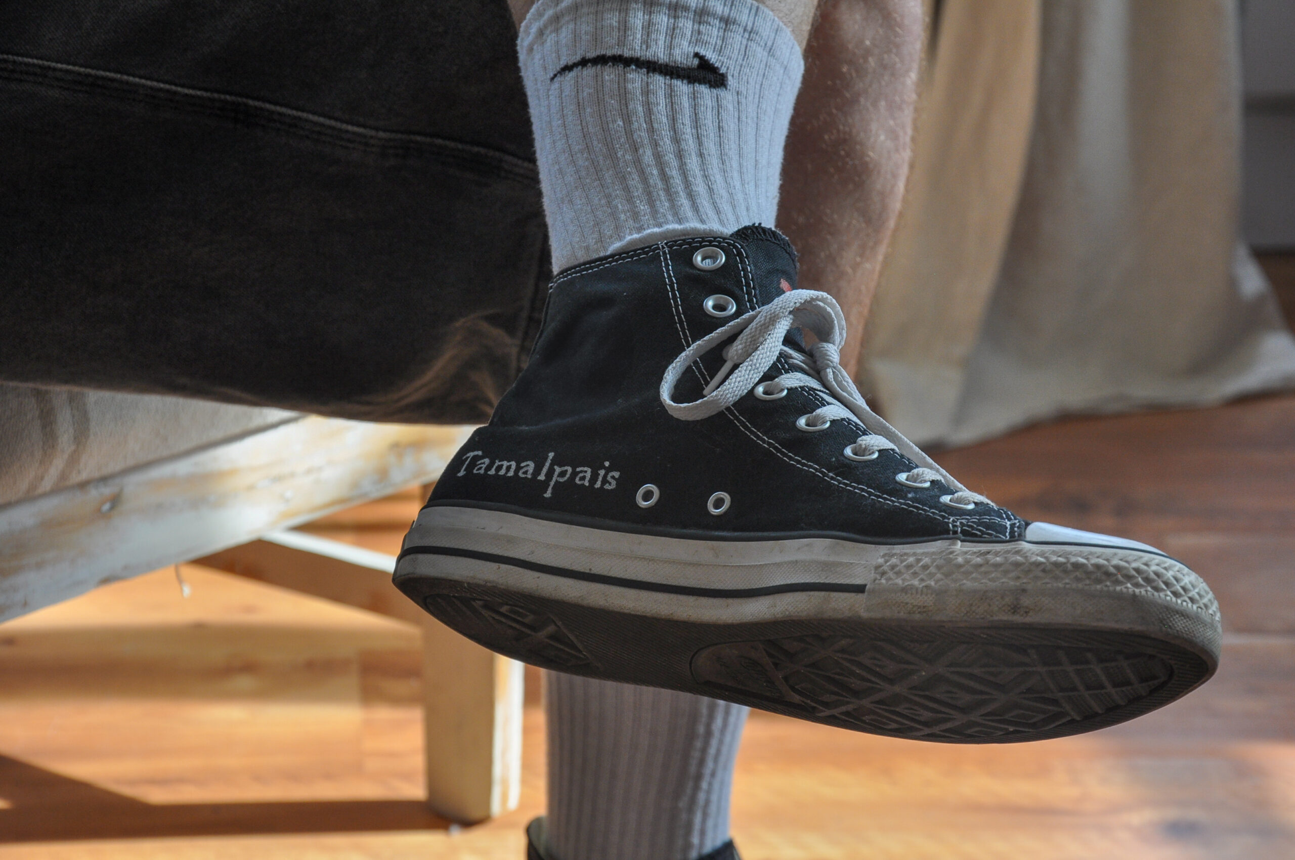 Close-up of a person wearing black Tamalpais high-top canvas sneakers with white laces, sitting on a chair indoors in natural light