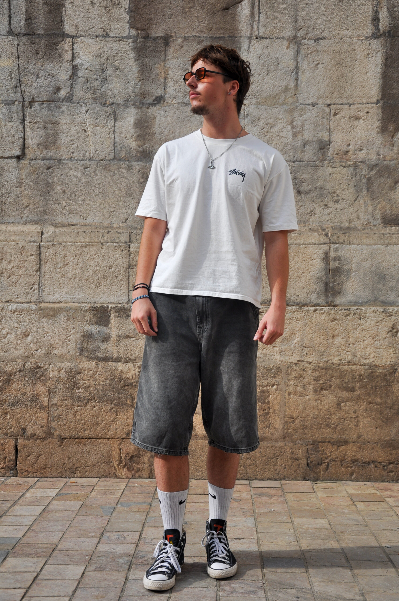 Young man standing against a textured stone wall, wearing a white Stüssy t-shirt, oversized black denim shorts, and black Tamalpais high-top sneakers with a bold logo on the tongue.
