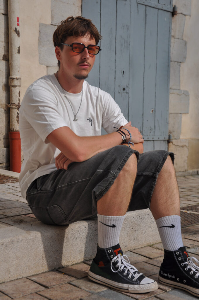 Young man sitting on stone steps wearing black Tamalpais high-top sneakers, white socks, oversized gray shorts, and a white t-shirt, posing in front of a rustic blue wooden door.