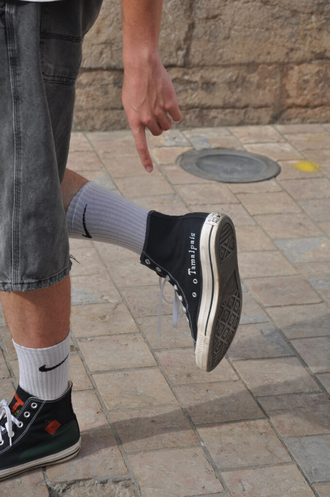 Close-up of a person walking on a stone pavement wearing black Tamalpais high-top sneakers with white laces, one foot lifted and pointing downward.